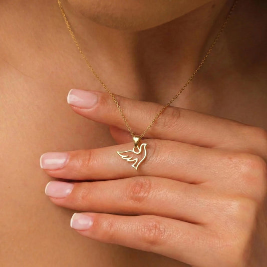 May include: A gold-colored necklace featuring a pendant in the shape of a dove with outstretched wings. The delicate chain and pendant are displayed against a skin-toned background. The dove pendant is a simple, elegant design.