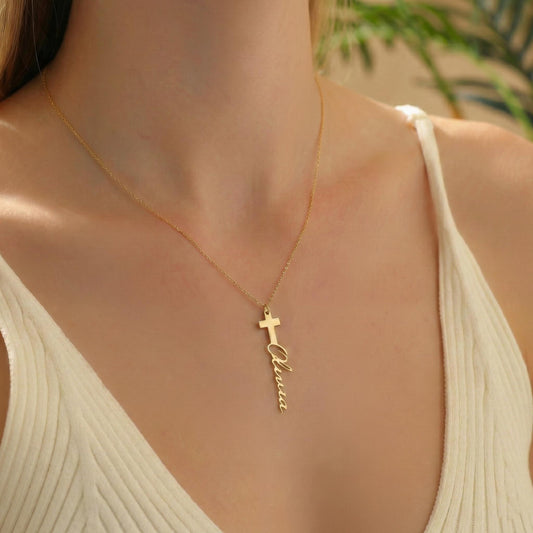 May include: A gold-colored necklace featuring a cross pendant with the name "Olivia" written vertically below it. The necklace is worn around the neck, with the chain visible against a white top.