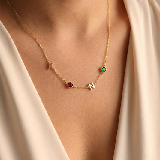 Dainty gold necklace featuring sideways initials and bezel set birthstones, personalized family jewelry for mother's day gift, minimalist custom letter necklace with ruby and emerald colored stones on neck.