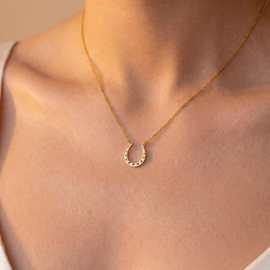 Dainty 14K gold horseshoe necklace worn delicately on a woman's neck, minimalist lucky charm pendant jewelry ideal for everyday wear and thoughtful Mother's day gift.