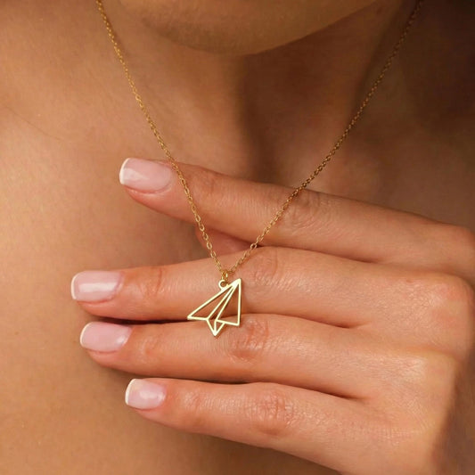 May include: A gold-colored necklace featuring a paper airplane pendant. The pendant is a minimalist design, with clean lines and a geometric shape. The necklace is worn around the neck.