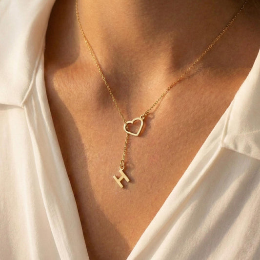 May include: A gold-colored necklace featuring a heart-shaped pendant and a dangling letter "H". The necklace is worn against a white shirt, creating a contrast that highlights the jewelry's design. The chain is delicate, and the pendant is simple yet elegant.