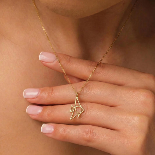 May include: A delicate gold-colored necklace featuring a pendant in the shape of a stylized fish. The pendant hangs from a fine chain, creating a minimalist and elegant design. The necklace is shown on a person's neck.