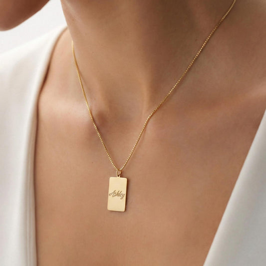 Gold vertical bar necklace on a model's neck, personalized with the engraved name Ashley in cursive font. Minimalist rectangular pendant gift for mother's day.