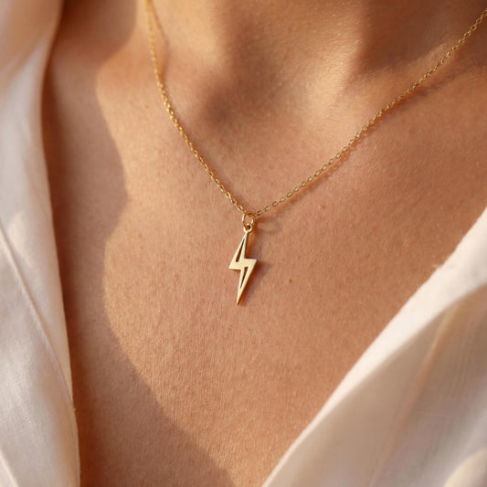 Close-up of a delicate minimalist 14k gold plated lightning bolt pendant necklace worn by a woman in a white shirt, glowing in soft sunlight.