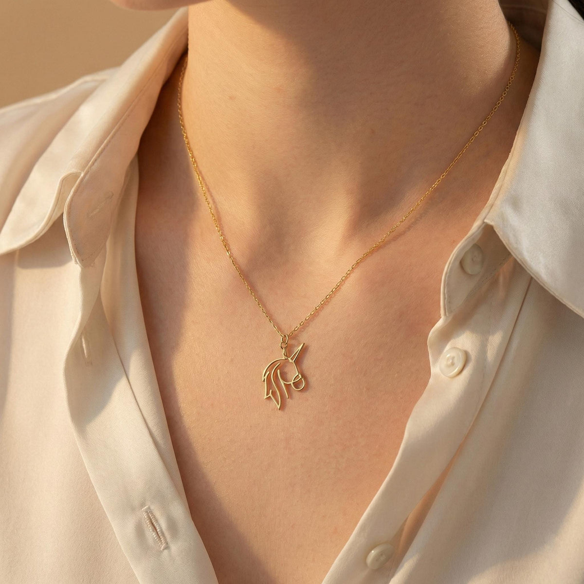 May include: A gold-colored necklace with a pendant shaped like a unicorn. The unicorn pendant is a minimalist design, featuring the outline of the mythical creature's head and horn. The necklace is worn against a white shirt.