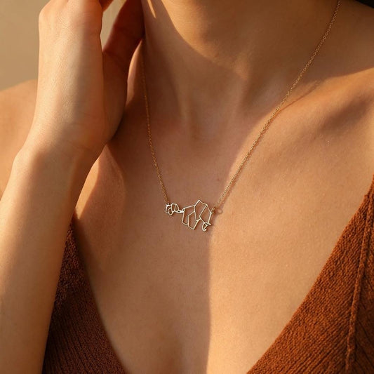 May include: A gold-colored necklace featuring a geometric elephant pendant. The pendant is a stylized representation of an elephant, with angular lines forming its shape. The necklace is worn around the neck.