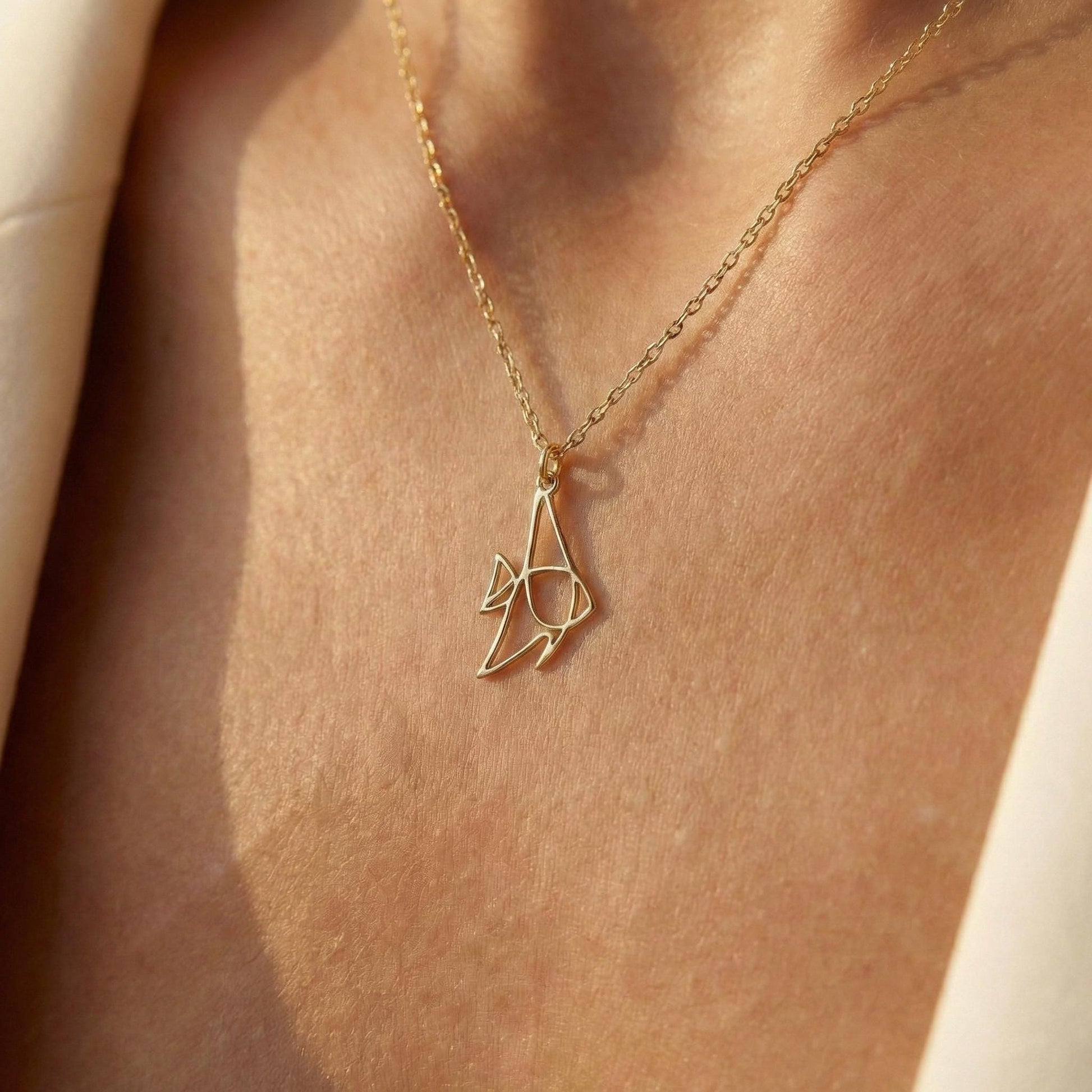 May include: A gold-colored pendant necklace featuring a stylized fish design. The pendant is suspended from a delicate chain. The necklace is worn against a skin tone background, highlighting the jewelry's design and color.