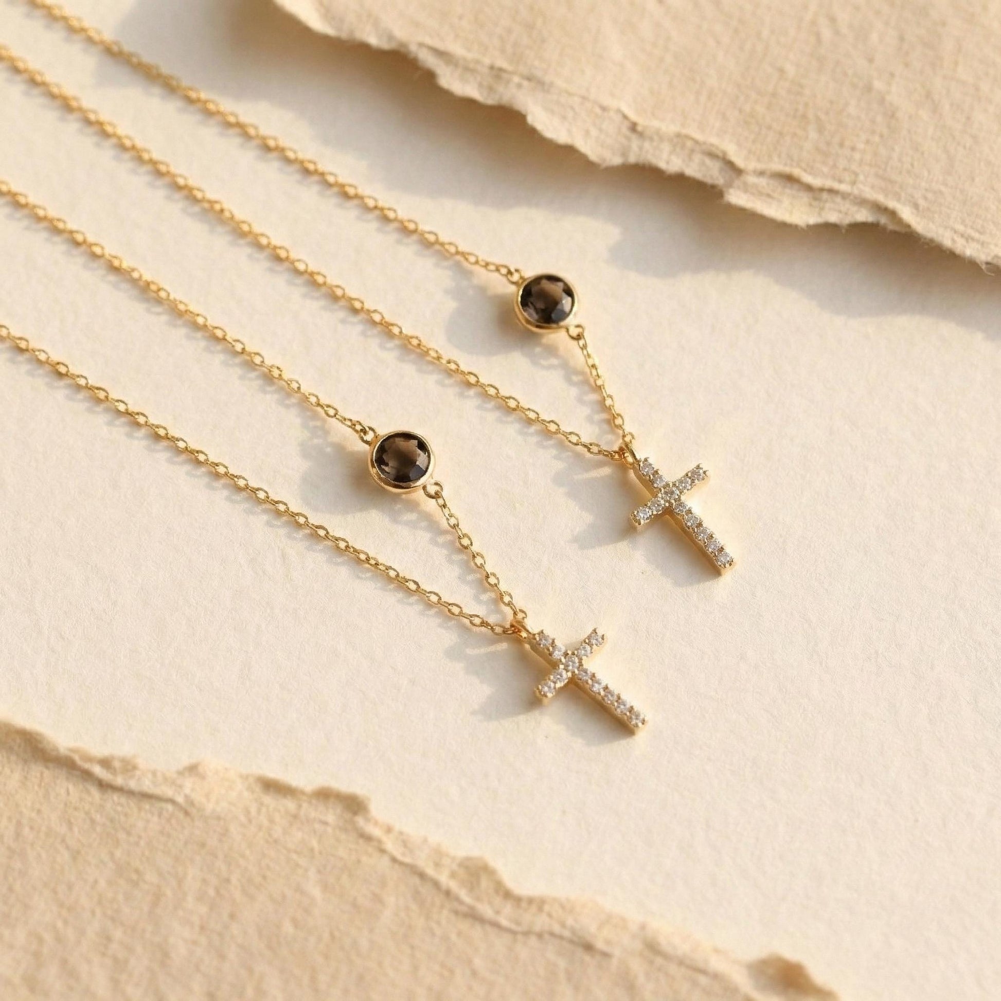 Dainty gold chain necklace featuring a small pave diamond cross pendant and a round bezel-set birthstone charm, worn by a model. Personalized minimalist religious jewelry.