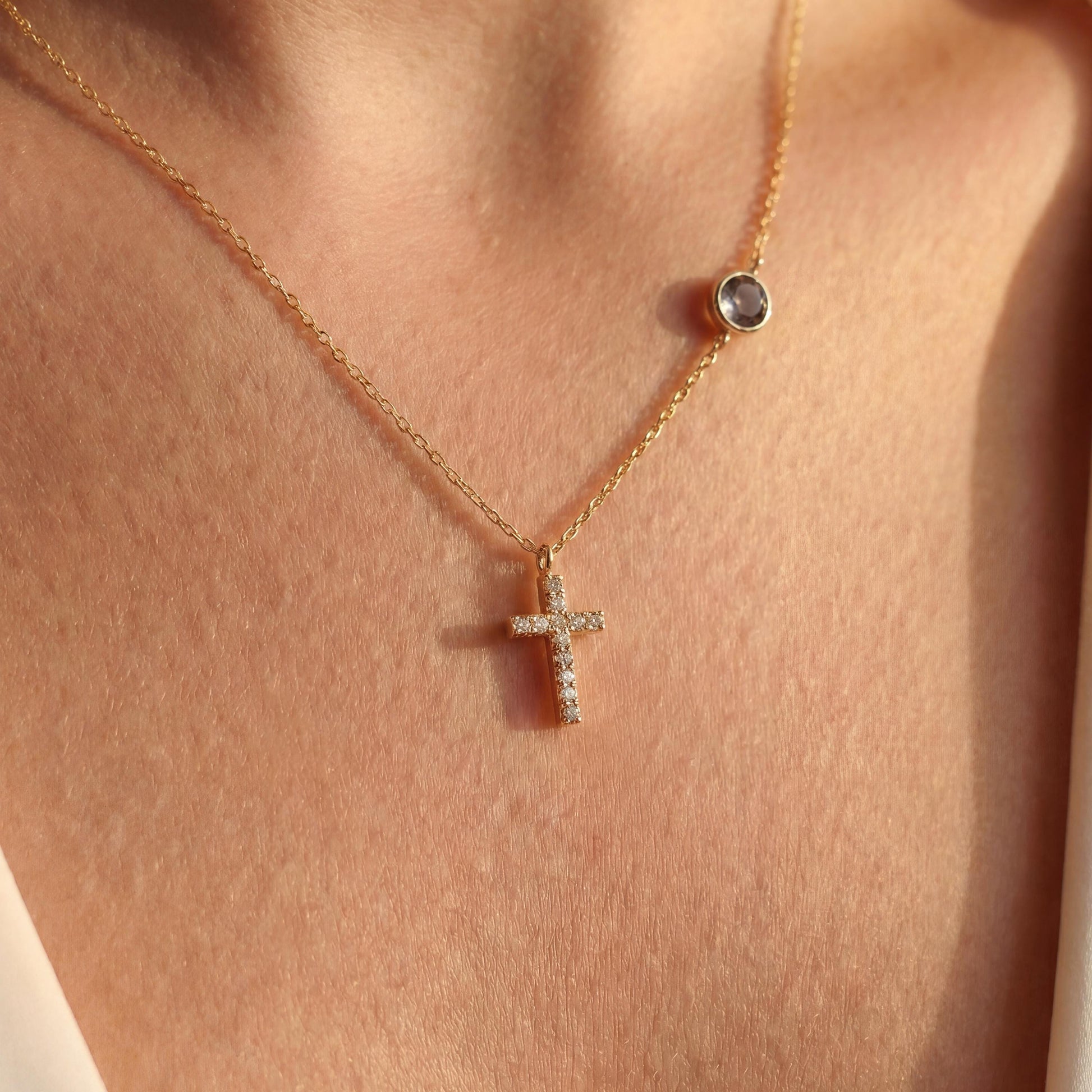 Dainty gold chain necklace featuring a small pave diamond cross pendant and a round bezel-set birthstone charm, worn by a model. Personalized minimalist religious jewelry.