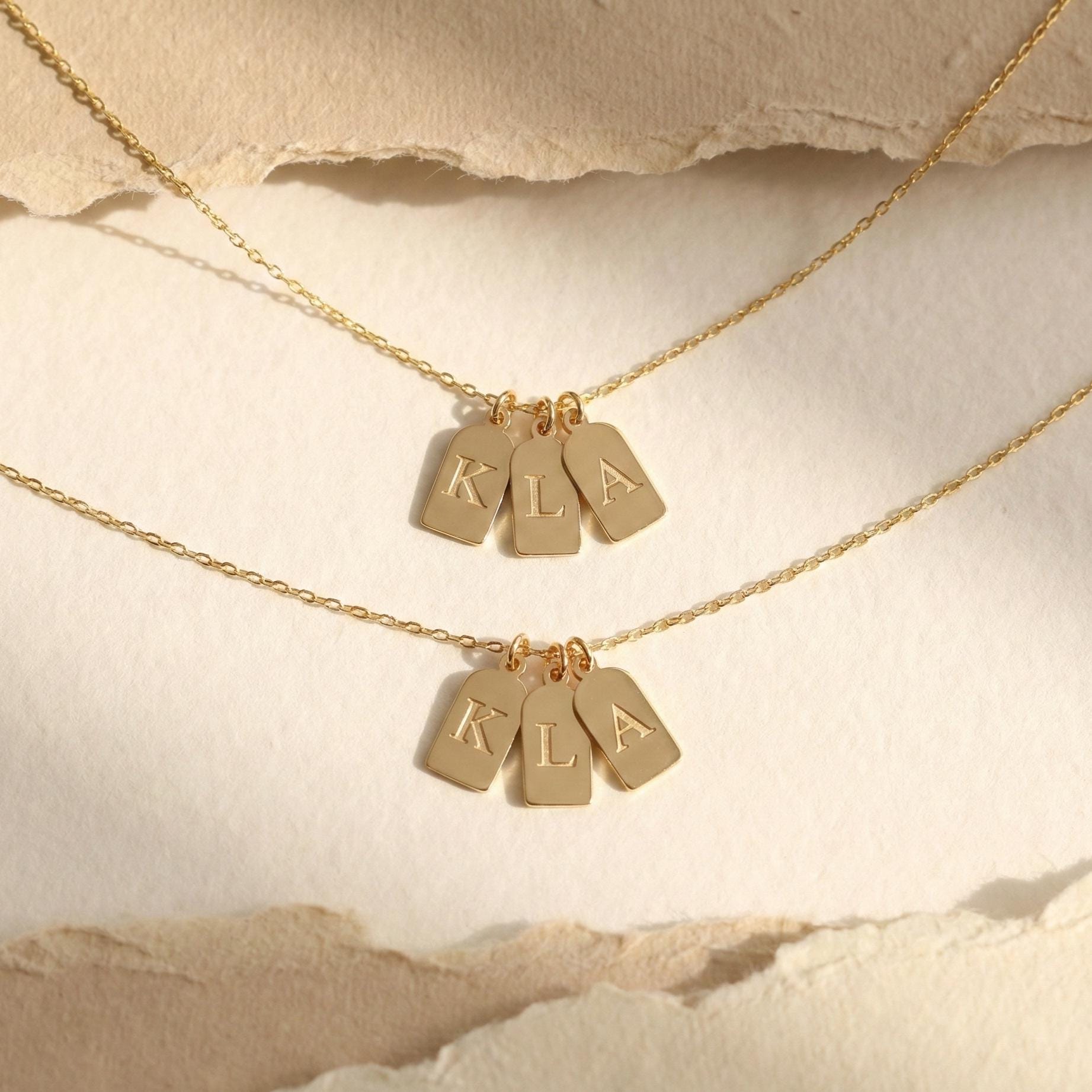 Gold initial necklace featuring three small rectangular pendant tags engraved with letters K L A, worn on a woman's neck, minimalist personalized jewelry for mom