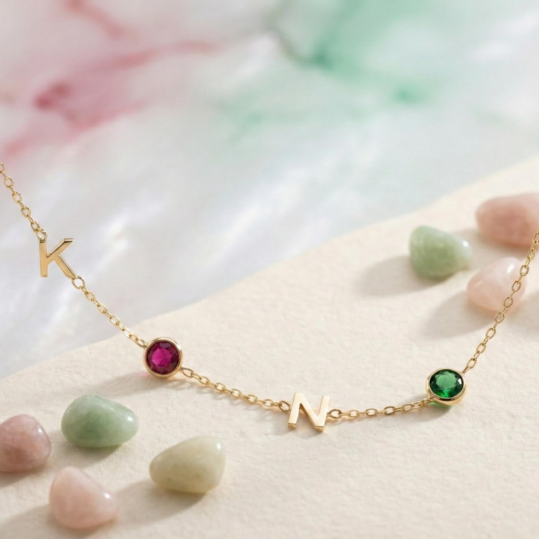 Dainty gold necklace featuring sideways initials and bezel set birthstones, personalized family jewelry for mother's day gift, minimalist custom letter necklace with ruby and emerald colored stones on neck.