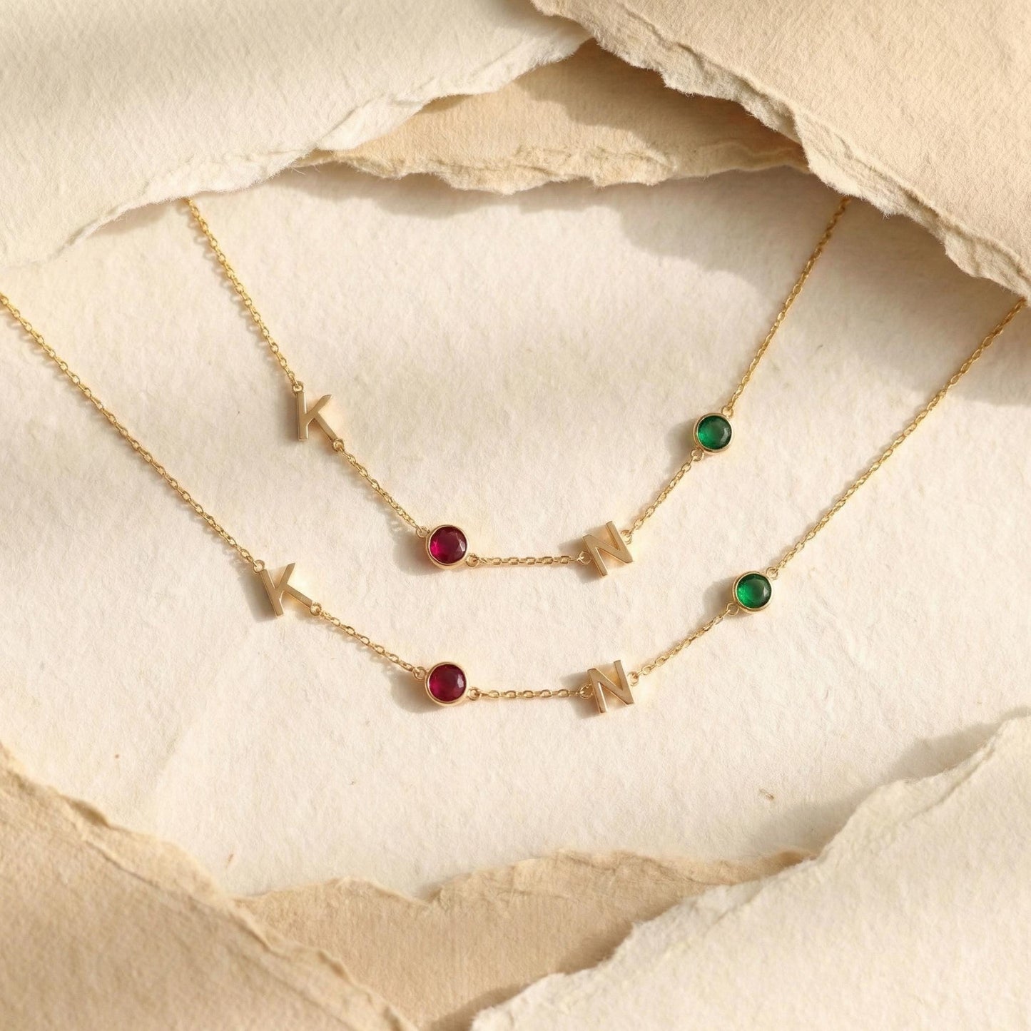Dainty gold necklace featuring sideways initials and bezel set birthstones, personalized family jewelry for mother's day gift, minimalist custom letter necklace with ruby and emerald colored stones on neck.