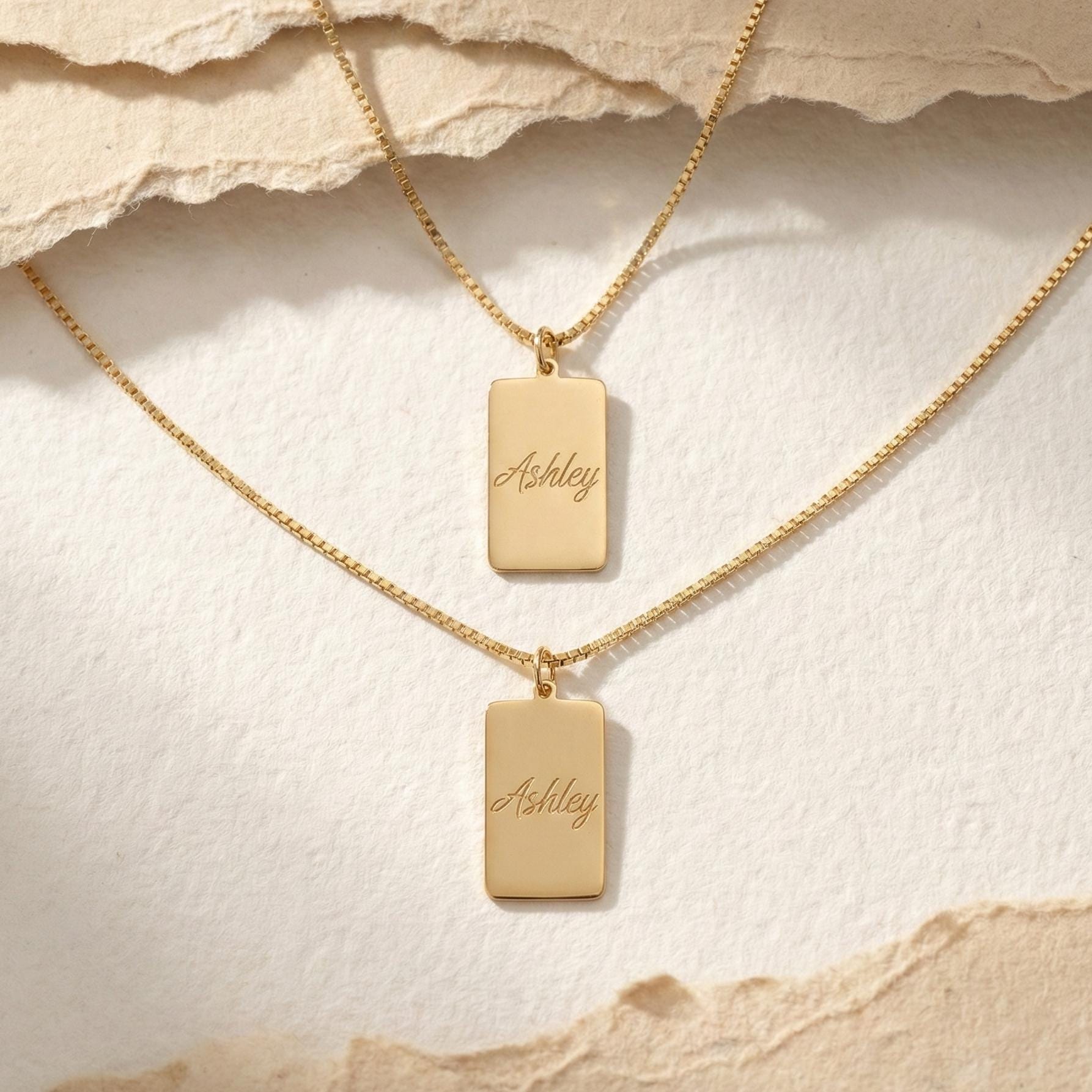 Gold vertical bar necklace on a model's neck, personalized with the engraved name Ashley in cursive font. Minimalist rectangular pendant gift for mother's day.