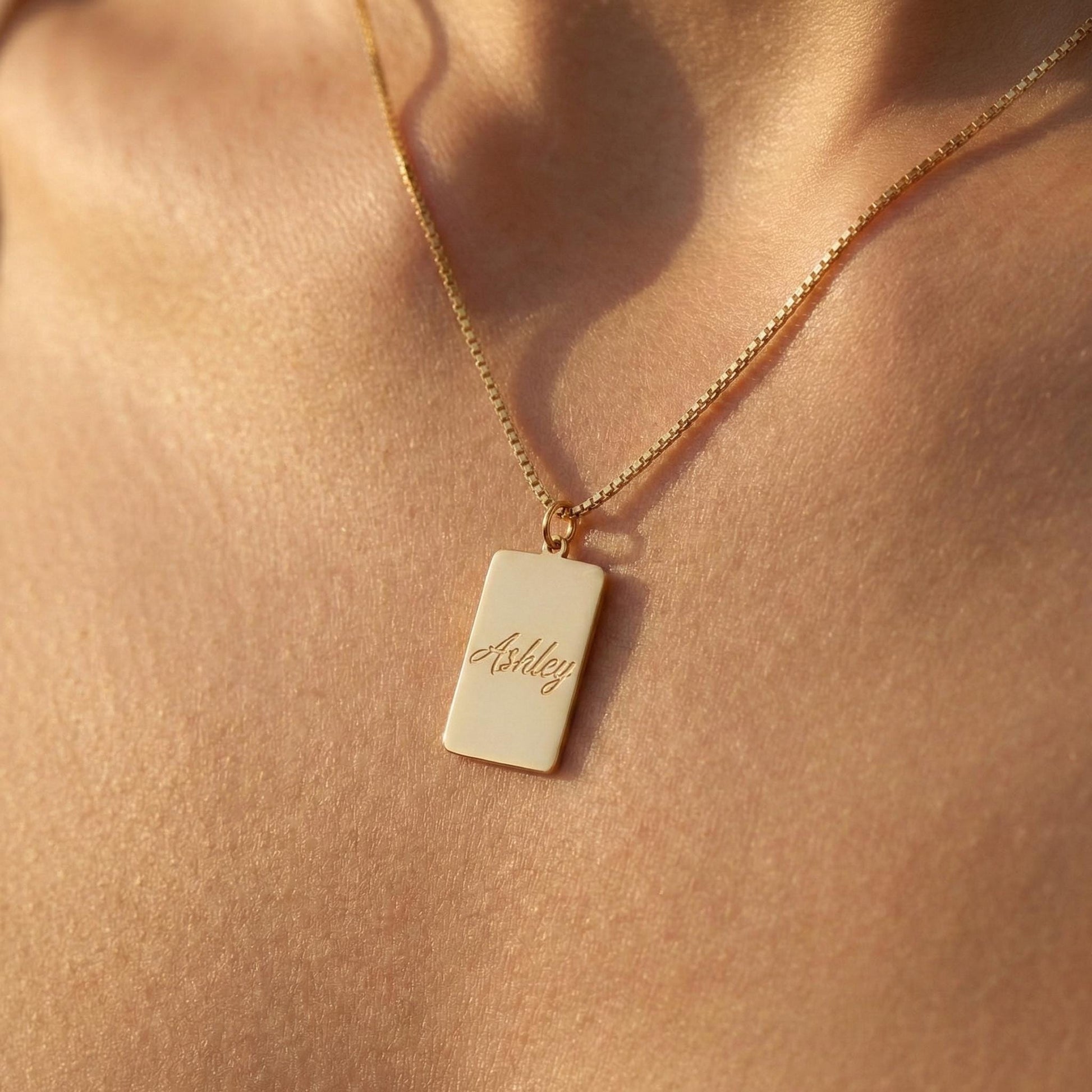 Gold vertical bar necklace on a model's neck, personalized with the engraved name Ashley in cursive font. Minimalist rectangular pendant gift for mother's day.