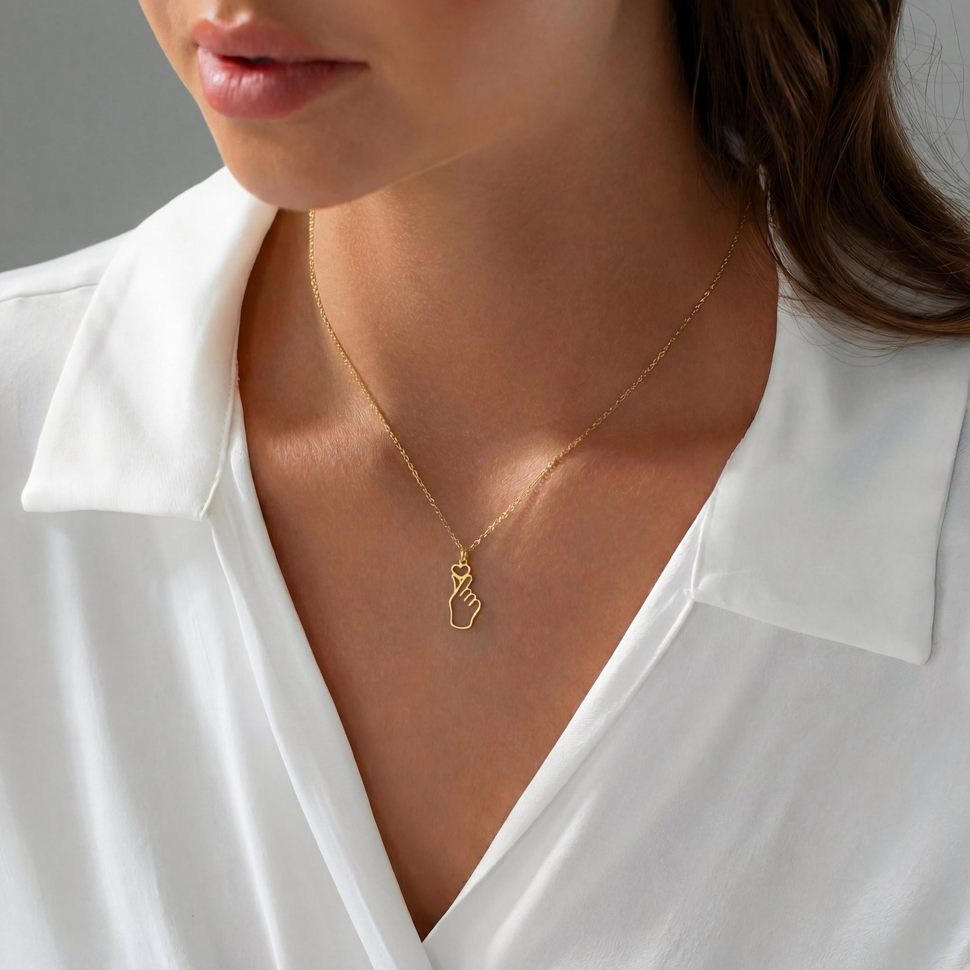 A woman wearing a dainty 14K gold minimalist Korean finger heart necklace, a delicate saranghae hand gesture pendant resting on her collarbone.