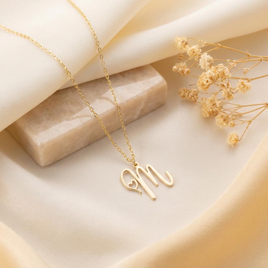 Dainty custom initial necklace with a delicate heart detail next to a cursive letter M, worn by a smiling woman in sunlight. Minimalist personalized elegant pendant.