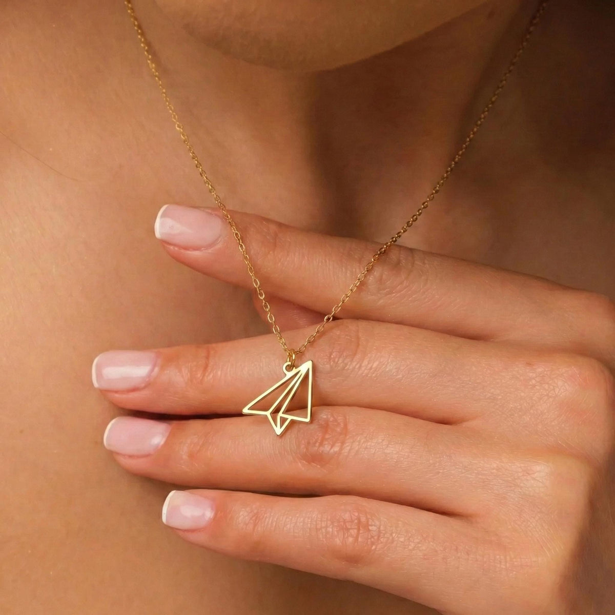 May include: A gold-colored necklace featuring a paper airplane pendant. The pendant is a minimalist design, with clean lines and a geometric shape. The necklace is worn around the neck.