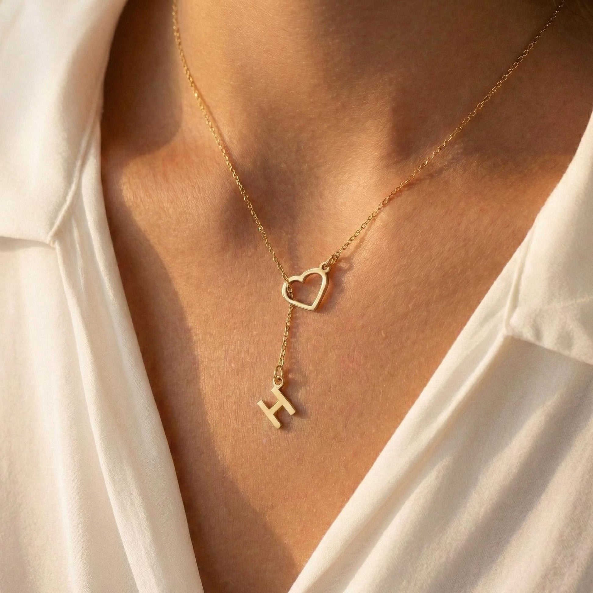 May include: A gold-colored necklace featuring a heart-shaped pendant and a dangling letter "H". The necklace is worn against a white shirt, creating a contrast that highlights the jewelry's design. The chain is delicate, and the pendant is simple yet elegant.