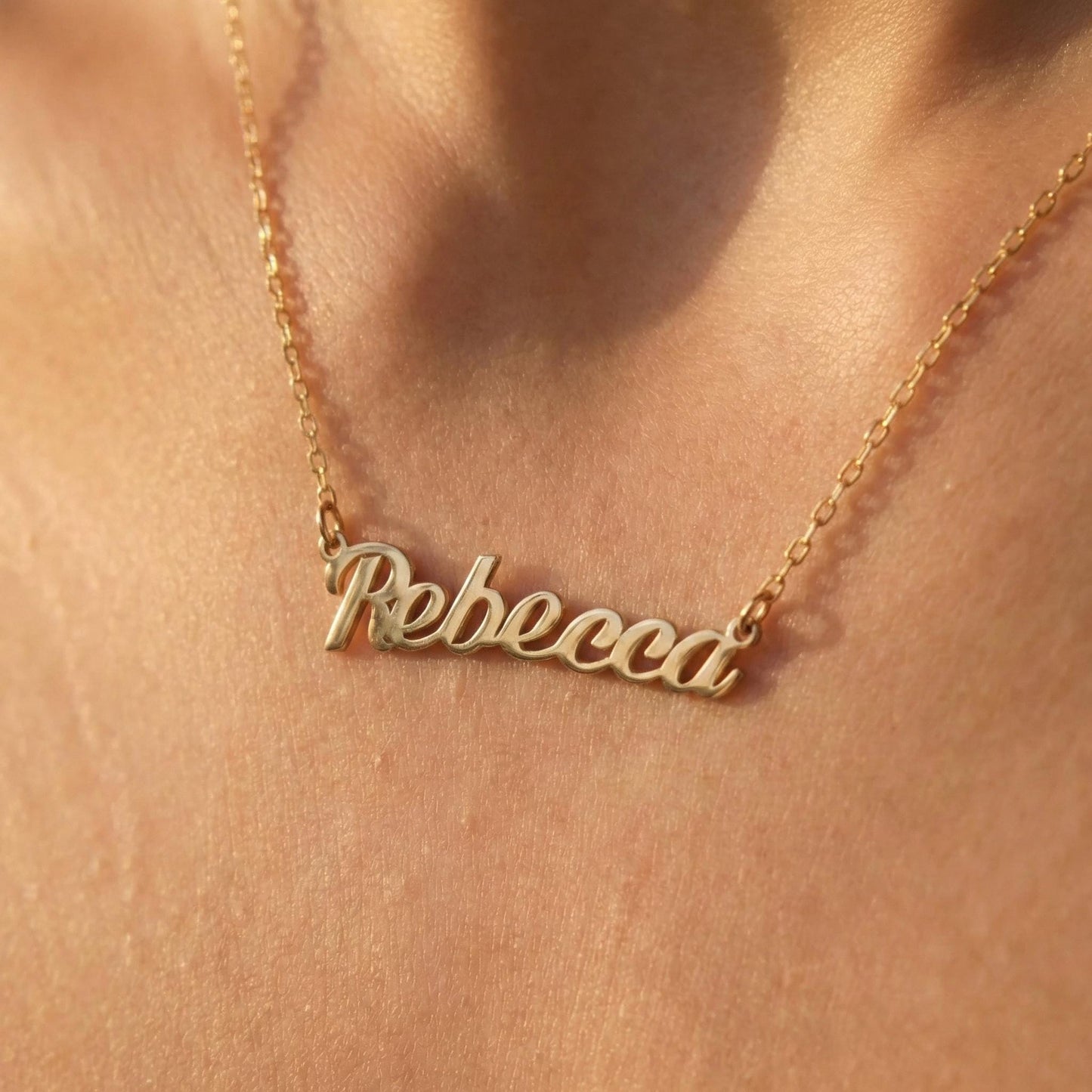 May include: A gold-colored necklace with the name "Rebecca" in cursive script. The pendant hangs from a delicate chain. The necklace is displayed against a skin-toned background, highlighting the jewelry's design.