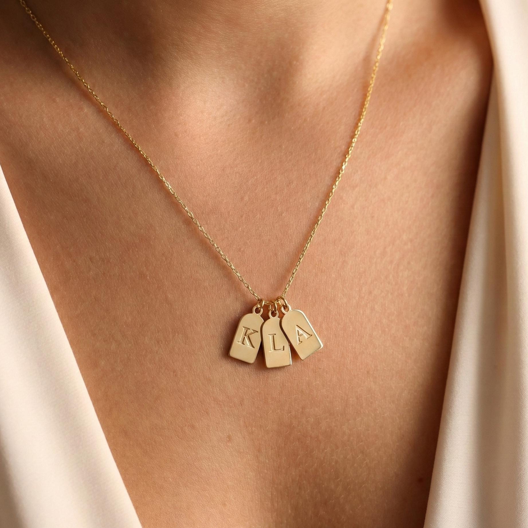 Gold initial necklace featuring three small rectangular pendant tags engraved with letters K L A, worn on a woman's neck, minimalist personalized jewelry for mom