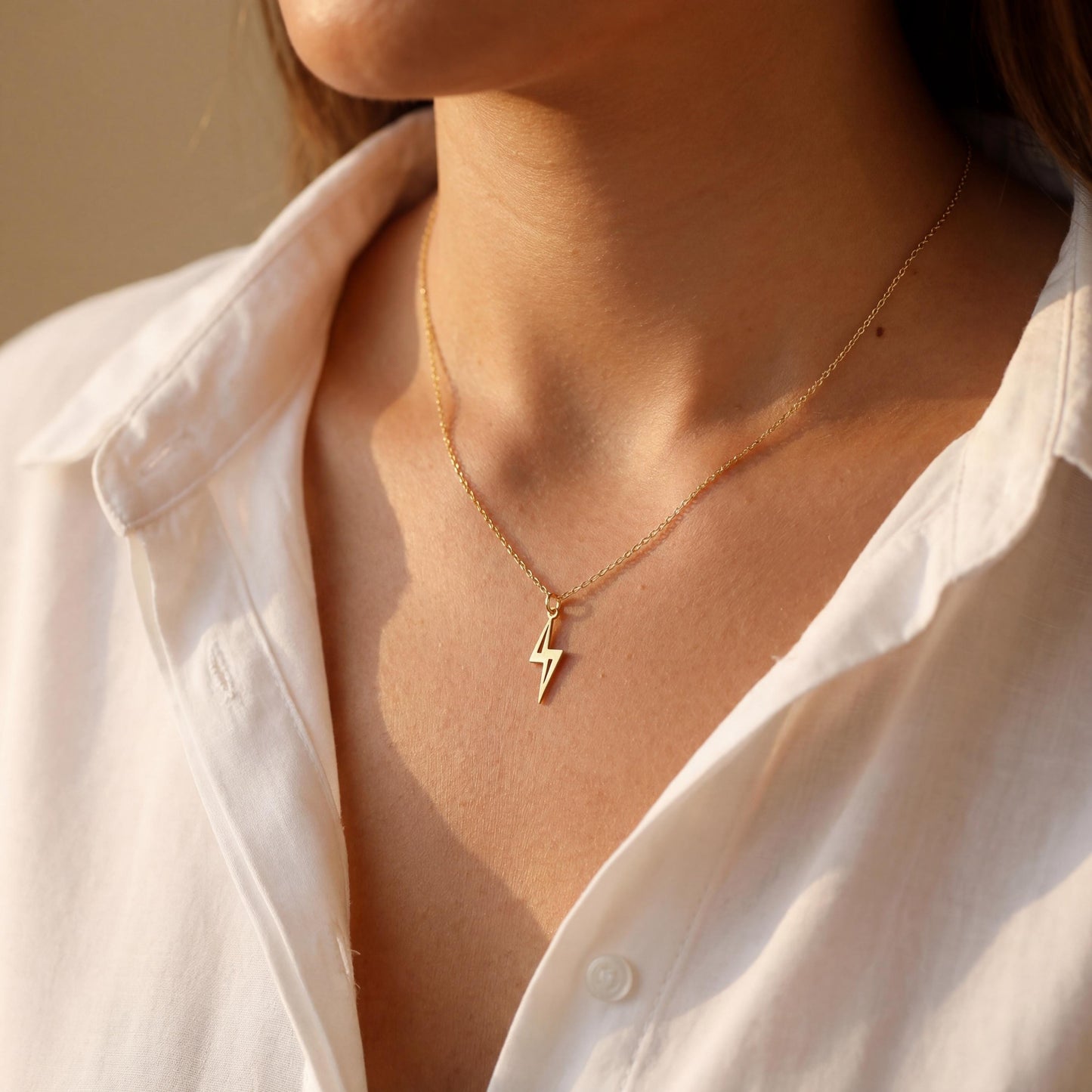Close-up of a delicate minimalist 14k gold plated lightning bolt pendant necklace worn by a woman in a white shirt, glowing in soft sunlight.
