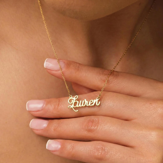 May include: A gold-colored necklace with the name "Lauren" in cursive script. The necklace is worn around the neck. The chain is delicate, and the name pendant is centered. The background is a person's skin.